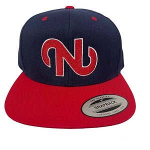 Puede incluir: Gorra snapback azul marino y roja con visera plana. La gorra presenta una gran letra "N" roja estilizada bordada en la parte delantera. La visera es roja y la corona es azul marino. Cierre a presi&oacute;n.
