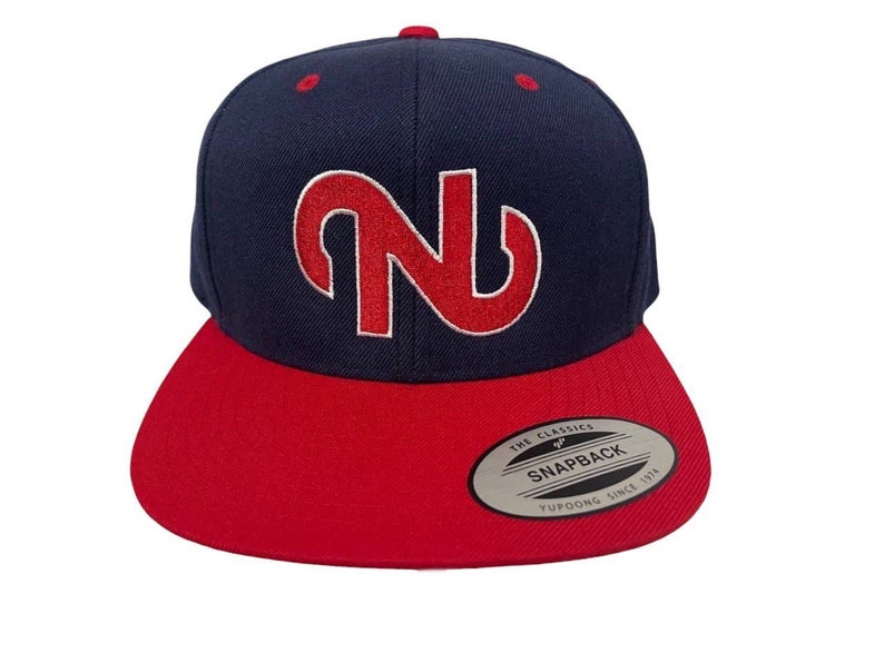 Puede incluir: Gorra snapback azul marino y roja con visera plana. La gorra presenta una gran letra "N" roja estilizada bordada en la parte delantera. La visera es roja y la corona es azul marino. Cierre a presi&oacute;n.