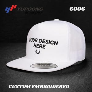 Może przedstawiać: Biała czapka typu snapback z siateczkowym tyłem i płaskim daszkiem. Na przednim panelu znajduje się czarny haftowany napis 'YOUR DESIGN HERE' nad małą ikoną wieńca laurowego. Na daszku znajduje się okrągła naklejka z napisem 'SNAPBACK'.