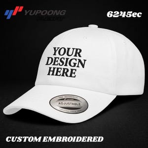 Può includere: Un cappellino da baseball bianco con il testo ricamato in nero YOUR DESIGN HERE sul pannello frontale. Il cappellino include un adesivo circolare argento e nero sulla visiera che recita THE AUTHENTIC YUPOONG ADJUSTABLE YUPOONG SINCE 1974.