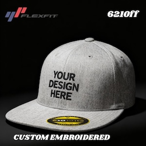 May include: A gray Flexfit baseball cap with a flat brim. The cap features the text "YOUR DESIGN HERE" embroidered in black. The words "CUSTOM EMBROIDERED" are at the bottom. The cap has a yellow and black sticker on the brim.