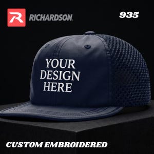 May include: A navy blue baseball cap with a mesh back and a flat brim. The front panel features white embroidered text that reads YOUR DESIGN HERE. The cap includes a top button, eyelets, and the Richardson logo with the model number 935.
