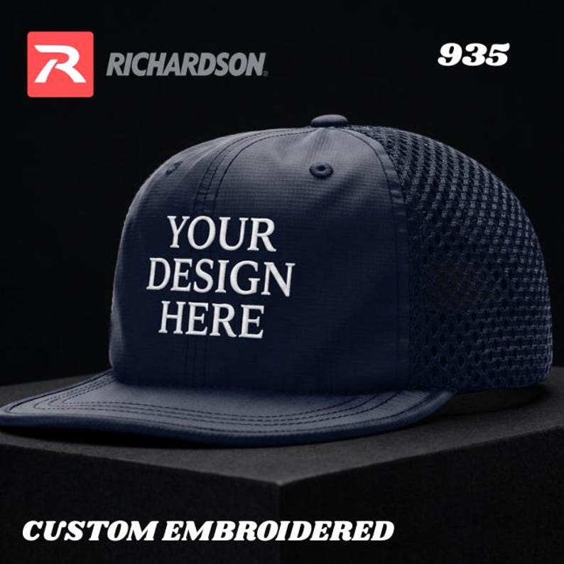 May include: A navy blue baseball cap with a mesh back and a flat brim. The front panel features white embroidered text that reads YOUR DESIGN HERE. The cap includes a top button, eyelets, and the Richardson logo with the model number 935.
