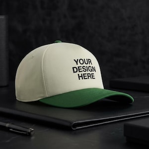 Puede incluir: Una gorra de béisbol con una corona de color crema y una visera de color verde oscuro. La parte frontal de la corona presenta el texto negro "YOUR DESIGN HERE" impreso en una fuente negrita sin remates. La gorra tiene un diseño estructurado.