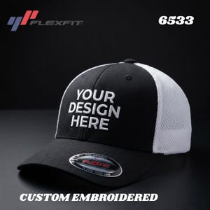 May include: Black and white Flexfit baseball cap with the text "YOUR DESIGN HERE" embroidered in white. The cap features a mesh back and a curved brim. The text "CUSTOM EMBROIDERED" is at the bottom.