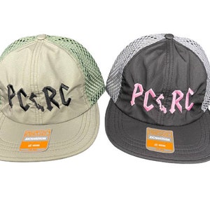 May include: Two trucker hats with mesh backs and flat brims. The left hat is beige with black embroidered text reading PC.RC. The right hat is dark gray with pink embroidered text reading PC.RC. Both hats feature a Richardson brand sticker on the brim.