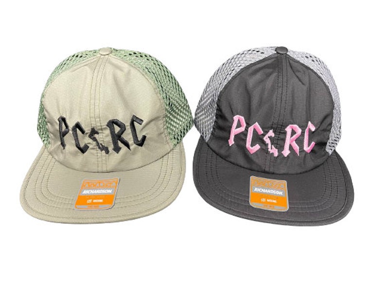 May include: Two trucker hats with mesh backs and flat brims. The left hat is beige with black embroidered text reading PC.RC. The right hat is dark gray with pink embroidered text reading PC.RC. Both hats feature a Richardson brand sticker on the brim.