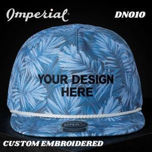 Op de afbeelding: Een op maat geborduurde baseballcap met een blauw tropisch bladerpatroon. De cap heeft de tekst "YOUR DESIGN HERE" in zwart, een wit touwdetail en de merknaam "Imperial".