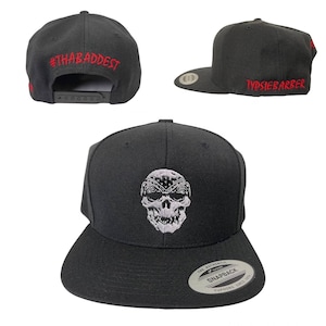 Puede incluir: Gorra snapback negra con dise&ntilde;o de calavera. El sombrero presenta una calavera bordada en blanco con un pa&ntilde;uelo. El texto "#THABADDEST" est&aacute; bordado en rojo en la parte posterior y "TYPSIEBARBER" en rojo en el lateral.