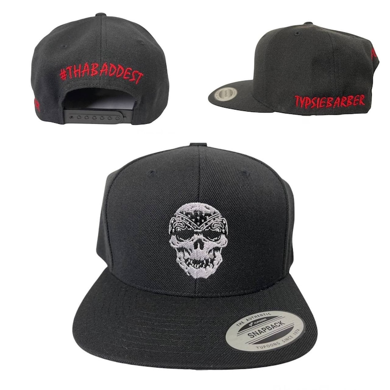 Puede incluir: Gorra snapback negra con dise&ntilde;o de calavera. El sombrero presenta una calavera bordada en blanco con un pa&ntilde;uelo. El texto "#THABADDEST" est&aacute; bordado en rojo en la parte posterior y "TYPSIEBARBER" en rojo en el lateral.