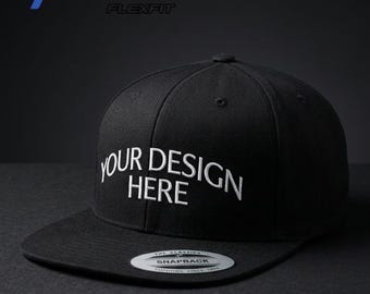 May include: A black Yupoong Flexfit 6089m snapback cap with a flat brim and embroidered white text on the front that reads YOUR DESIGN HERE. The cap features a circular silver and black sticker on the brim and includes the brand logo in the corner.