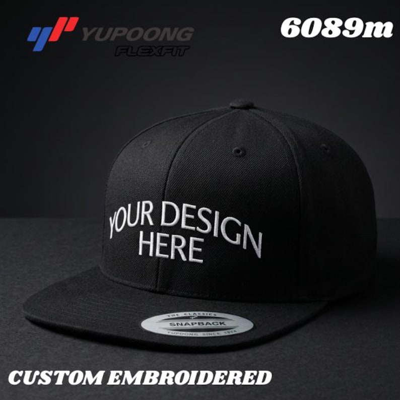 Puede incluir: Gorra de b&eacute;isbol negra Flexfit con el texto "YOUR DESIGN HERE" bordado en blanco. La gorra tiene una visera plana y un cierre snapback. Las palabras "CUSTOM EMBROIDERED" est&aacute;n en la parte inferior.