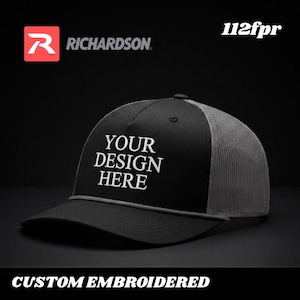 Puede incluir: Gorra de camionero negra y gris con el texto "YOUR DESIGN HERE" en blanco. La gorra tiene una visera curva y una parte trasera de malla. El logotipo de Richardson es visible en el lado izquierdo. La parte inferior de la imagen dice "CUSTOM EMBROIDERED".