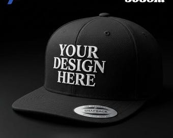 May include: A black Yupoong 5089m snapback cap featuring the white embroidered text YOUR DESIGN HERE on the front panel. The hat has a flat brim with a circular silver and black sticker that reads THE CLASSIC SNAPBACK SNAPBACK ONLY YUPOONG.