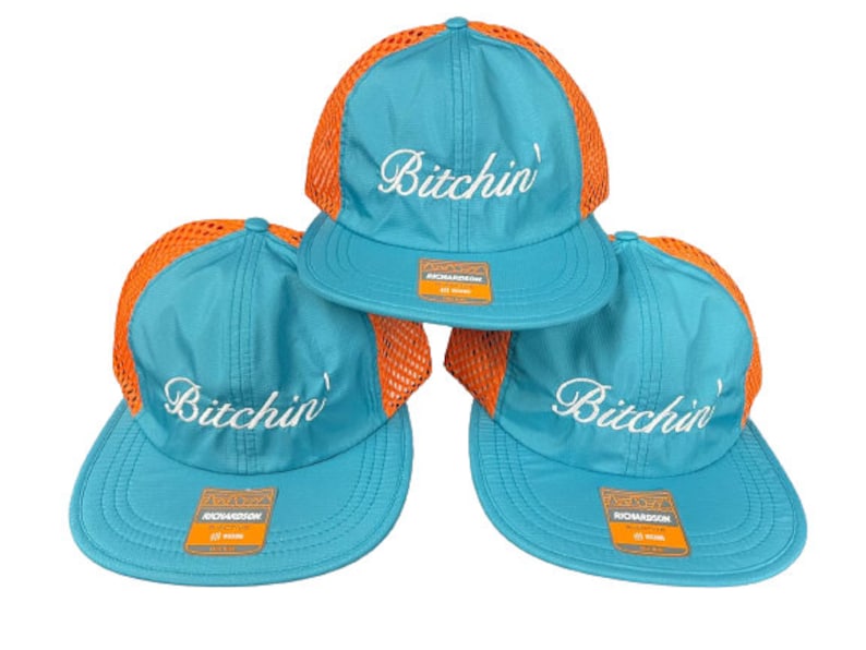 May include: Three turquoise baseball caps with orange mesh back panels. Each cap features the word Bitchin' embroidered in white script on the front crown and a rectangular Richardson brand sticker on the flat brim of each hat.