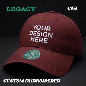 May include: A maroon baseball cap with the text "YOUR DESIGN HERE" embroidered in white. The cap has a curved brim and a small circular tag. The words "LEGACY", "CFA", and "CUSTOM EMBROIDERED" are also visible.