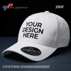 May include: White Flexfit Delta baseball cap with the text "YOUR DESIGN HERE" embroidered in black. The cap has a curved brim and a Flexfit Delta label. The words "CUSTOM EMBROIDERED" are at the bottom.