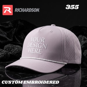 May include: A light purple five-panel baseball cap with a white rope detail across the base of the crown. The front features the embroidered text "YOUR DESIGN HERE" in white. The side panels have laser-cut ventilation holes.