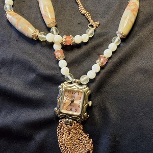 May include: A gold chain necklace with a square watch pendant. The watch has a pink face with black numbers and a gold tassel. The necklace is made of white and pink beads with brown and white striped beads.