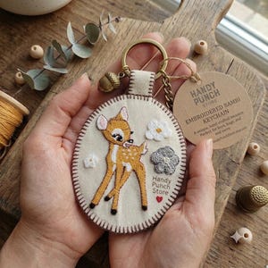 May include: An oval-shaped embroidered keyring featuring a fawn with white spots on a cream-coloured fabric background. The design includes small white and grey flowers, a tiny red heart, and the text Handy Punch Store. It has a gold metal ring.