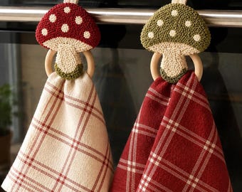 May include: Two hanging kitchen towels with mushroom-shaped toppers. One towel is cream with red plaid, the other is red with white plaid. The toppers are made of textured yarn in red and green with white dots, attached to wooden rings.