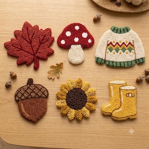 Puede incluir: Un conjunto de seis posavasos de punch needle con una hoja de arce roja, una seta roja y blanca, un suéter crema con patrón geométrico, una bellota marrón, un girasol amarillo y un par de botas de lluvia amarillas sobre una superficie de madera.
