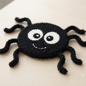 May include: A round, black crocheted spider coaster featuring two large white circular eyes with black pupils and a small white stitched smile. The spider has eight curved, textured black crocheted legs extending from its circular body.