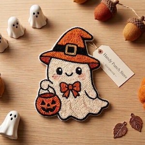 May include: A punch needle ghost decoration wearing an orange witch hat and a bow tie, holding an orange jack-o'-lantern bucket. The ghost has a tag labelled Handy Punch Store. Surrounding items include small white ghost figures, felt acorns, and a pumpkin.