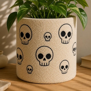 May include: A cylindrical woven plant pot cover in a cream color featuring a repeating pattern of embroidered black skulls in various sizes. The textured fabric has a dense, knotted appearance, and a green leafy plant is placed inside the container.