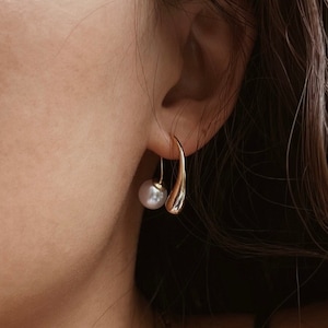 May include: A woman wearing a gold necklace and gold earrings with a pearl accent. The earrings are a curved shape.