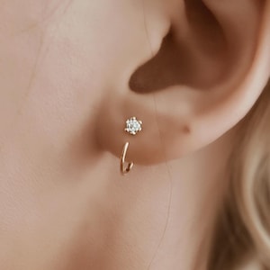 Puede incluir: Primer plano de un pendiente dorado con una pequeña gema brillante. El pendiente tiene un diseño delicado, con un pequeño aro que se curva hacia arriba hasta una sola piedra transparente. El pendiente se lleva en una oreja, con la piel y el pelo visibles.