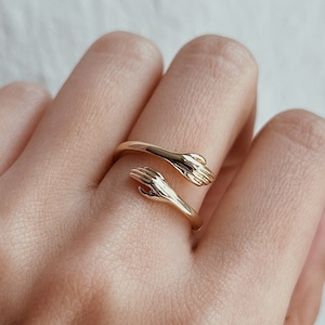 Op de afbeelding: Gouden ring met twee handen die in elkaar grijpen en een cirkel vormen. De ring wordt gedragen aan een vinger van een hand met een zwarte polkadotmouw.