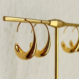 Op de afbeelding: Een paar gouden creolen met een gebogen, golvend ontwerp. De oorbellen worden gepresenteerd op een gouden standaard.