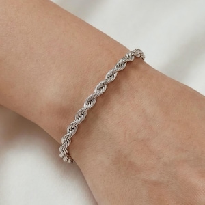 Könnte beinhalten: Ein silbernes Armband mit Seilkette. Das Armband besteht aus eng verdrehten Metallgliedern, die eine strukturierte Oberfläche bilden. Es wird an einem Handgelenk vor einem weißen Hintergrund getragen.