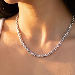 Könnte beinhalten: Eine silberfarbene Halskette mit Seilkette. Die Kette besteht aus einer Reihe von verdrehten Gliedern, die ein seilartiges Aussehen erzeugen. Die Kette wird um den Hals getragen und liegt auf dem Schlüsselbein. Der Hintergrund hat einen weichen, warmen Ton.