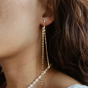 May include: A woman wearing a white top with a pearl necklace and long gold chain earrings.