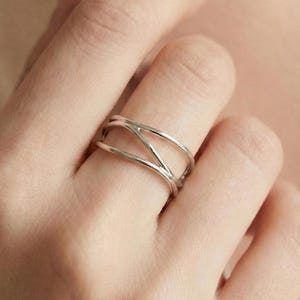 Puede incluir: Un anillo de plata con un diseño abierto único. El anillo presenta múltiples bandas que se cruzan y crean un patrón geométrico llamativo. El anillo se lleva en un dedo, mostrando su estilo elegante y moderno.