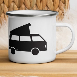 May include: White enamel mug with a black graphic of a camper van. The mug has a silver rim and a white handle. The mug is sitting on a wooden tray with cookies nearby. The background includes a woven basket and white fur.