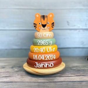 Könnte beinhalten: Ein hölzernes Stapelspielzeug mit einem Tigerkopf als Abschluss und fünf Ringen in den Farben Beige, Grün, Gelb, Orange und Braun. Die Ringe sind mit dem Text 50 cm, 2965 g, 20:40 Uhr, 18.04.2024 und dem Namen Janno in weißer Schrift bedruckt.