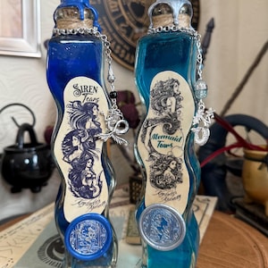 Op de afbeelding: Twee glazen flessen met blauwe en turquoise vloeistof. De flessen zijn gelabeld met "Siren Tears" en "Mermaid Tears". De flessen hebben zilveren en blauwe waszegels.
