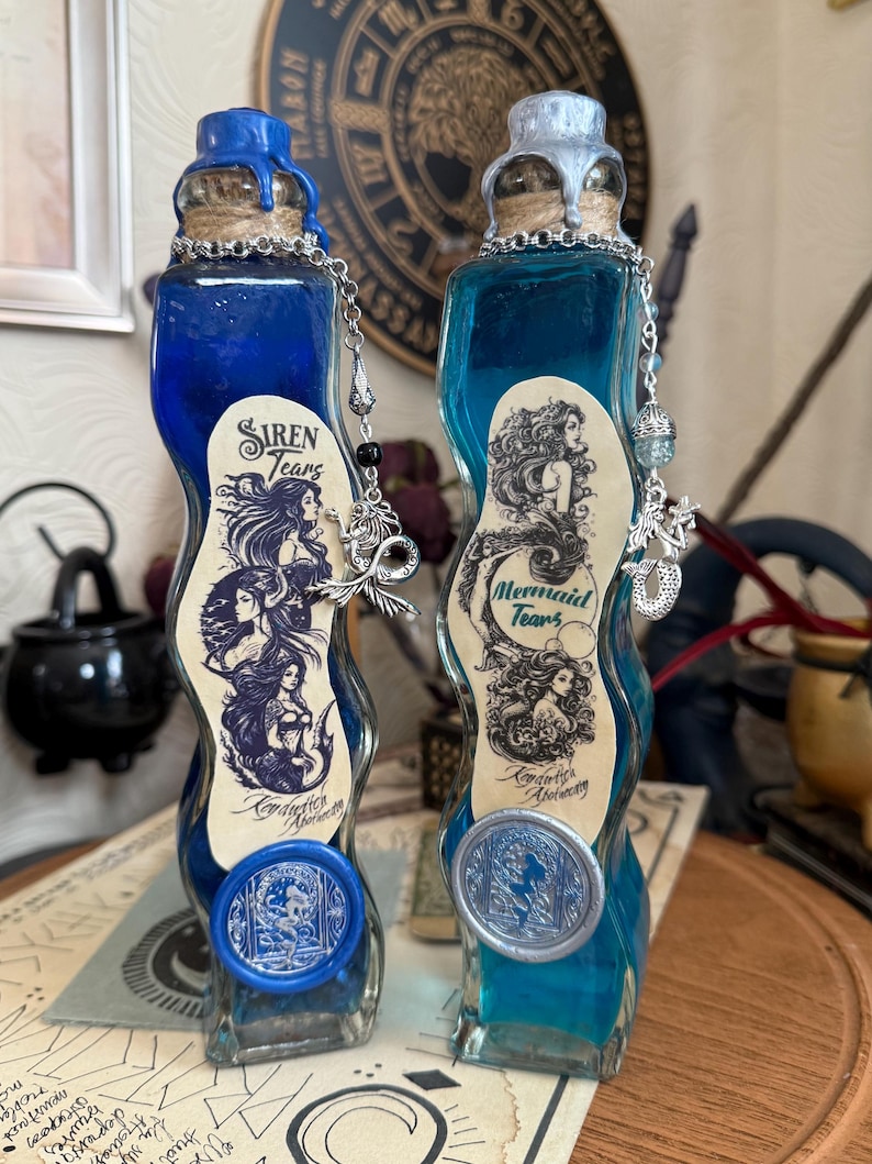 Op de afbeelding: Twee glazen flessen met blauwe en turquoise vloeistof. De flessen zijn gelabeld met "Siren Tears" en "Mermaid Tears". De flessen hebben zilveren en blauwe waszegels.