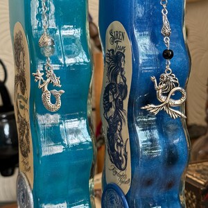 Op de afbeelding: Twee blauwe glazen flessen met zilveren zeemeermincharms en etiketten met de tekst "Mermaid Tears" en "Siren Tears". De flessen hebben een golvend, getextureerd ontwerp en zijn gevuld met een glinsterende blauwe vloeistof.