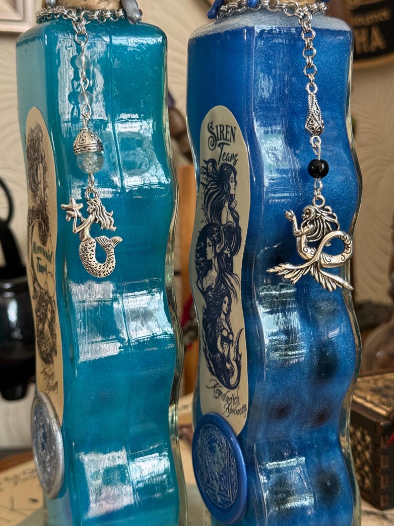 Op de afbeelding: Twee blauwe glazen flessen met zilveren zeemeermincharms en etiketten met de tekst "Mermaid Tears" en "Siren Tears". De flessen hebben een golvend, getextureerd ontwerp en zijn gevuld met een glinsterende blauwe vloeistof.