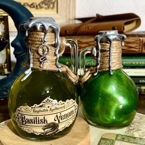 May include: Two small glass bottles with green liquid inside. The bottles have silver lids with a wax-like drip effect and are wrapped in twine. The bottle on the left has a label that reads "Keydwitch Apothecary Basilisk Venom".