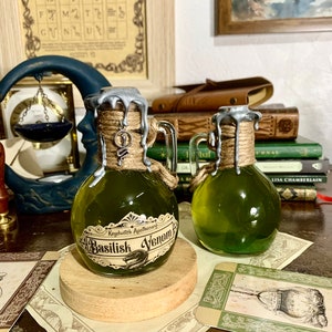 May include: Two small glass bottles with green liquid and silver lids. The bottles are labeled "Keydwitch Apothecary Basilisk Venom". The bottles are sitting on a wooden surface with a brown background.