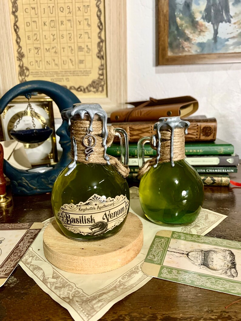 May include: Two small glass bottles with green liquid and silver lids. The bottles are labeled "Keydwitch Apothecary Basilisk Venom". The bottles are sitting on a wooden surface with a brown background.
