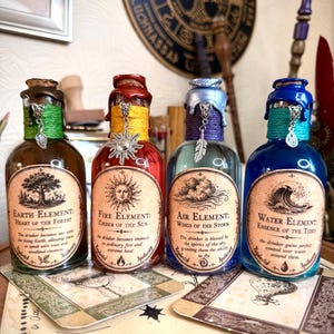 May include: Four glass bottles labelled Earth Element, Fire Element, Air Element, and Water Element. Each bottle features a cork stopper sealed with coloured wax, a decorative metal charm, and a coloured twine wrap around the neck of the bottle.