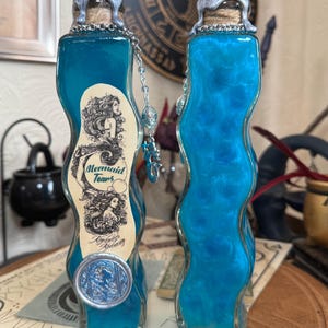 Op de afbeelding: Twee blauwe glazen flessen met zilveren doppen en kurken. De flessen zijn gevuld met een blauwe vloeistof en hebben een etiket met de tekst "Mermaid Tears, London Alchemy".