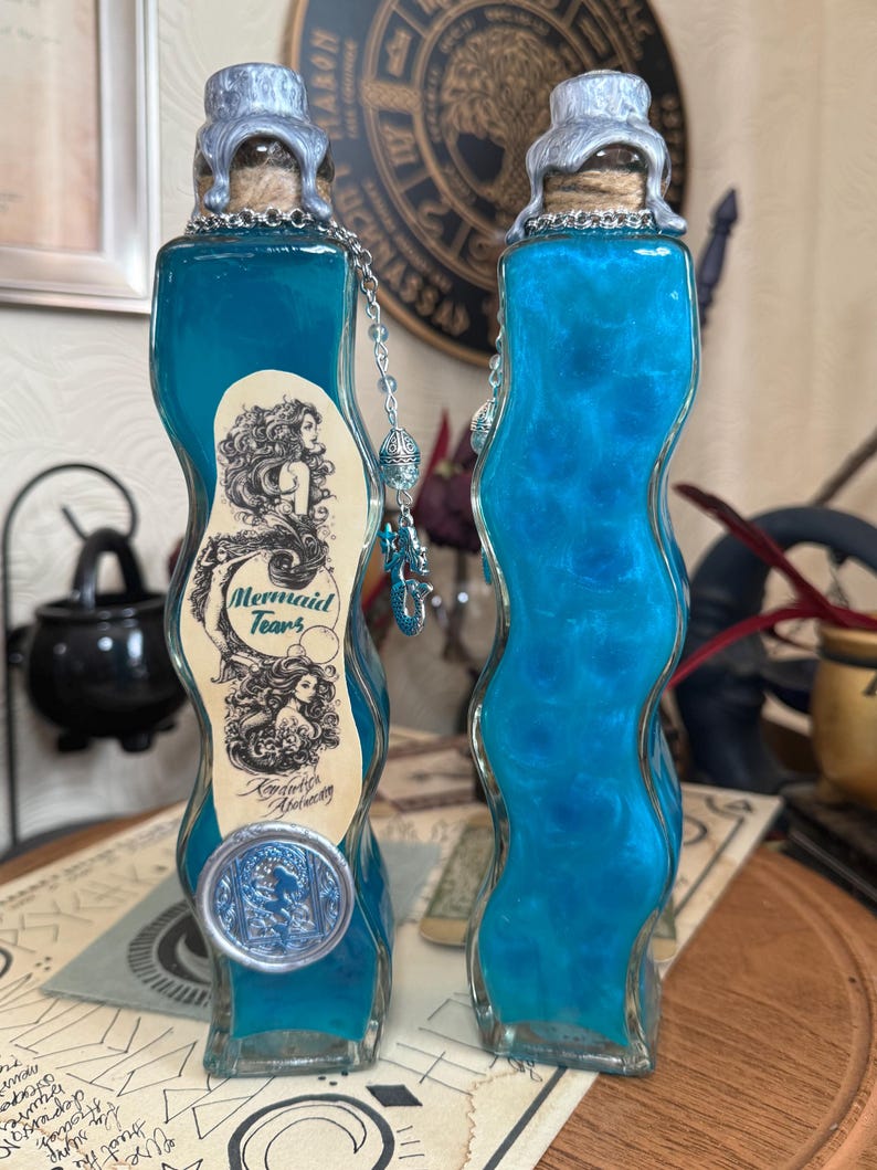 Op de afbeelding: Twee blauwe glazen flessen met zilveren doppen en kurken. De flessen zijn gevuld met een blauwe vloeistof en hebben een etiket met de tekst "Mermaid Tears, London Alchemy".