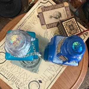 Op de afbeelding: Twee decoratieve glazen flessen met blauwe en turquoise vloeistof. De flessen hebben iriserende flesstoppen met zeemeerminschubben en zilveren kettingen.